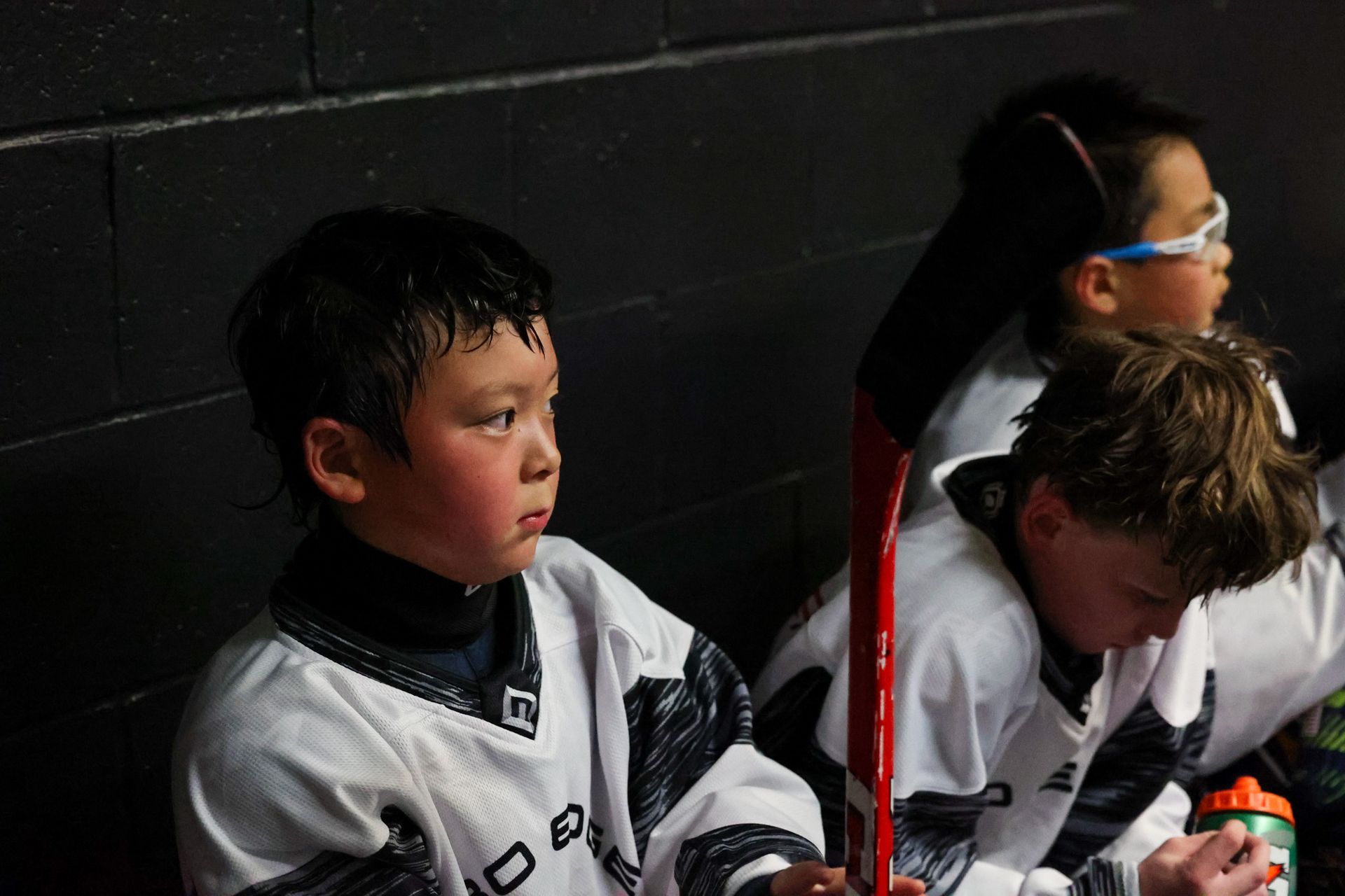 Three youth hockey players in white jerseys sit on the bench, one holding a red stick.