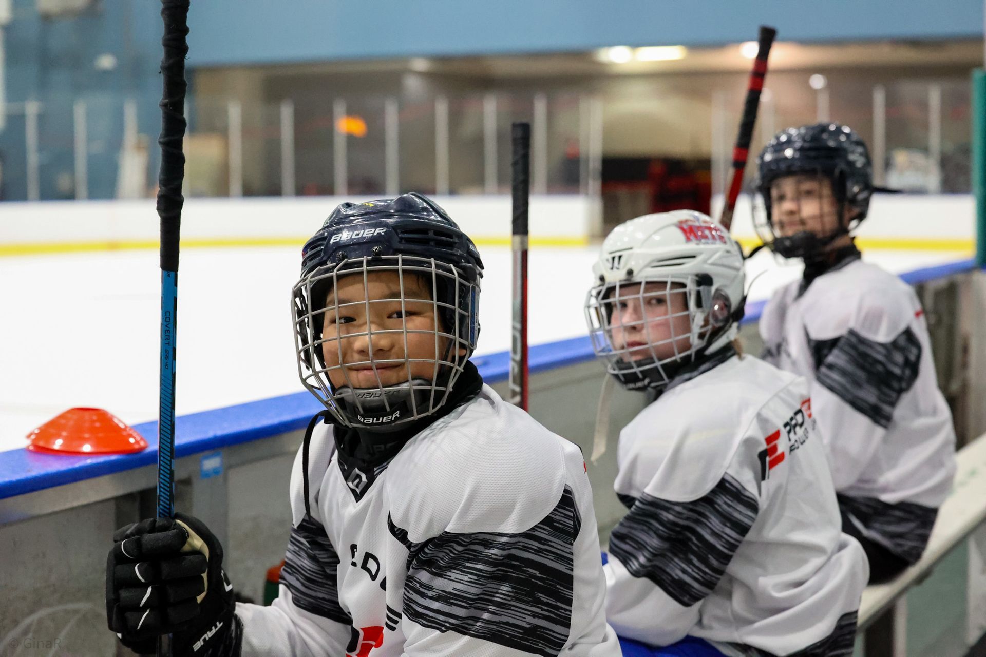 Three youth hockey players in white jerseys sit on a rink bench, wearing helmets and holding sticks.