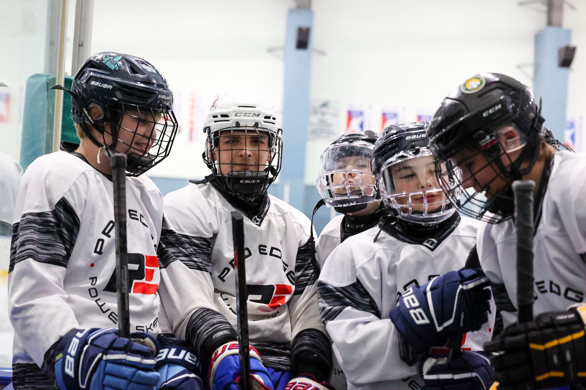 Youth ice hockey team huddled on the rink in white jerseys and helmets, listening closely.