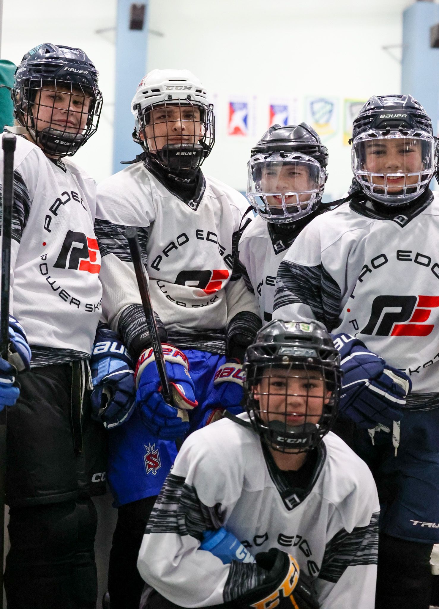 Youth hockey team in white jerseys posing in a hallway, some holding sticks and wearing blue or black gear