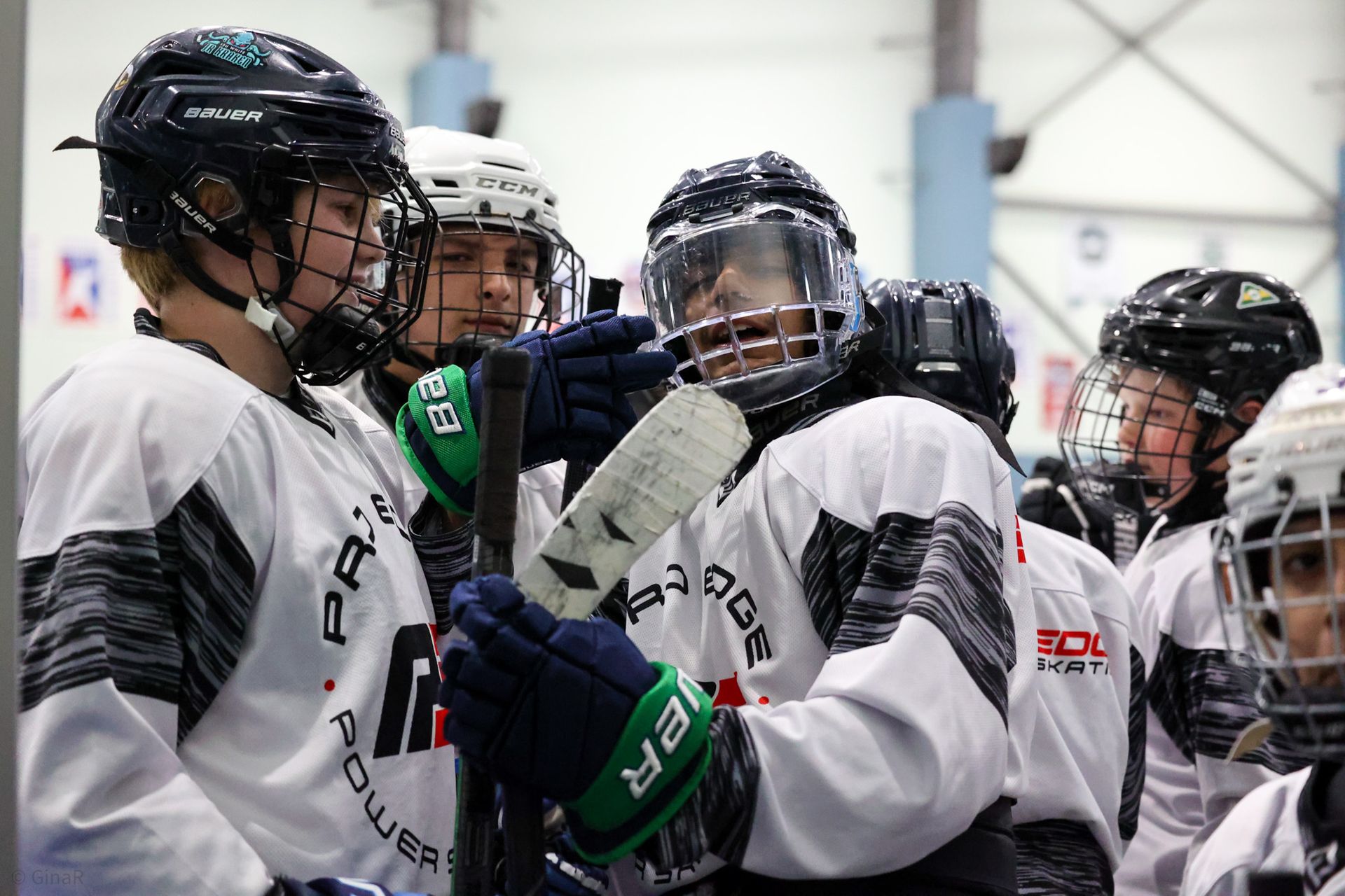 Ice hockey players in white jerseys huddle together on the rink, wearing helmets and gloves.