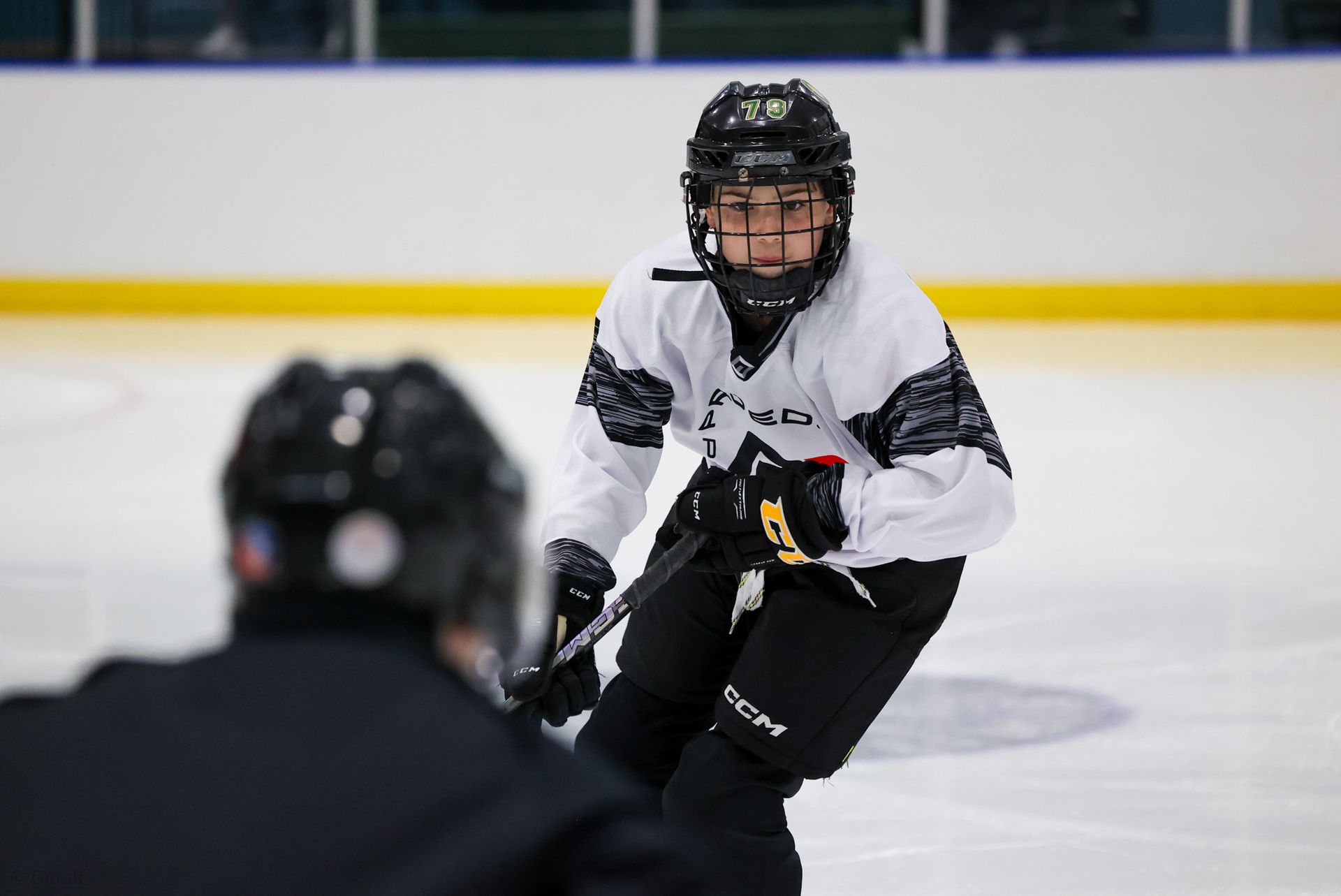 Hockey player in white jersey skating toward the camera on an ice rink, with a blurred player in foreground