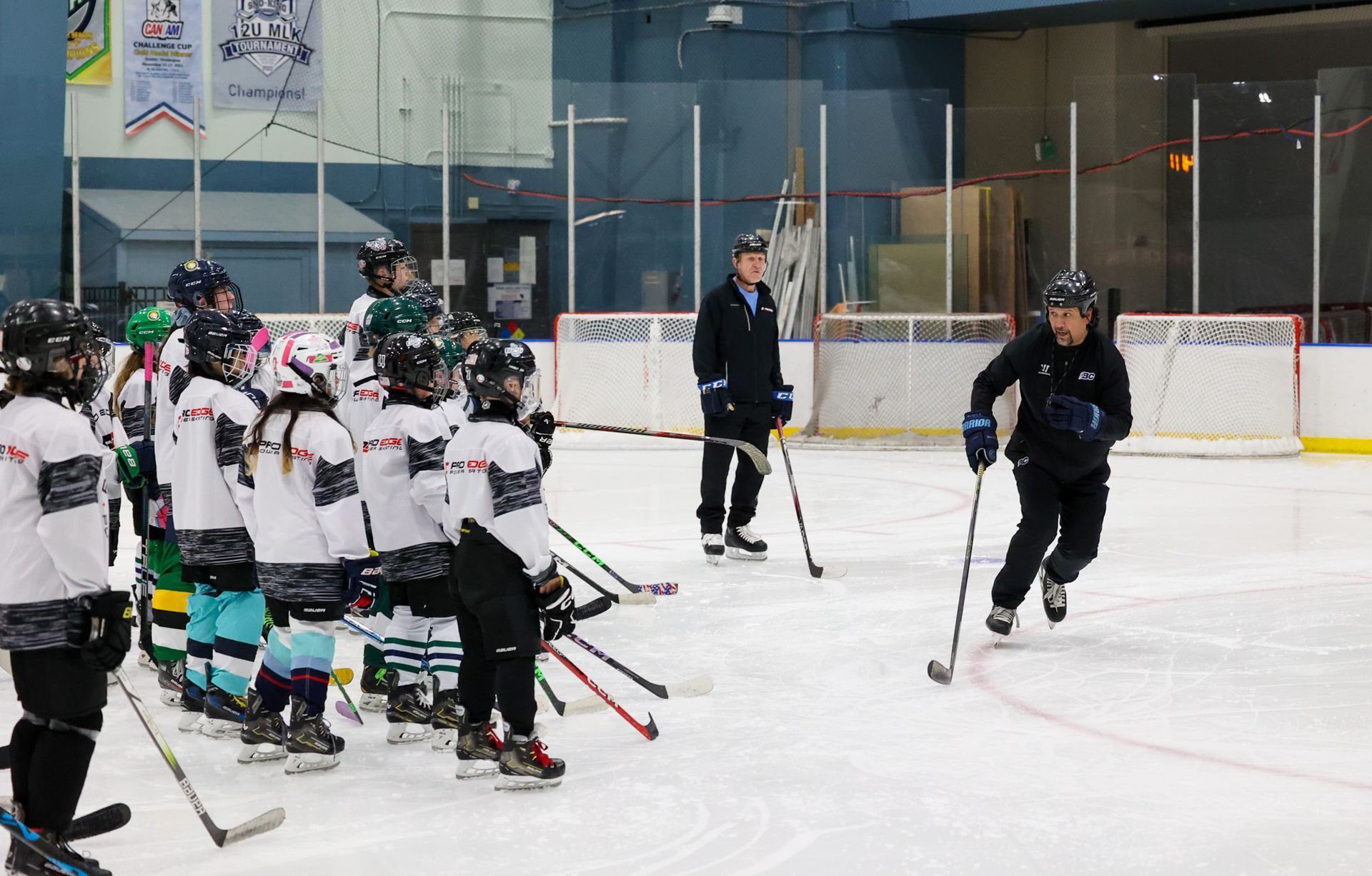 Youth hockey practice on an indoor rink, with a coach and players in white jerseys gathered near the boards.