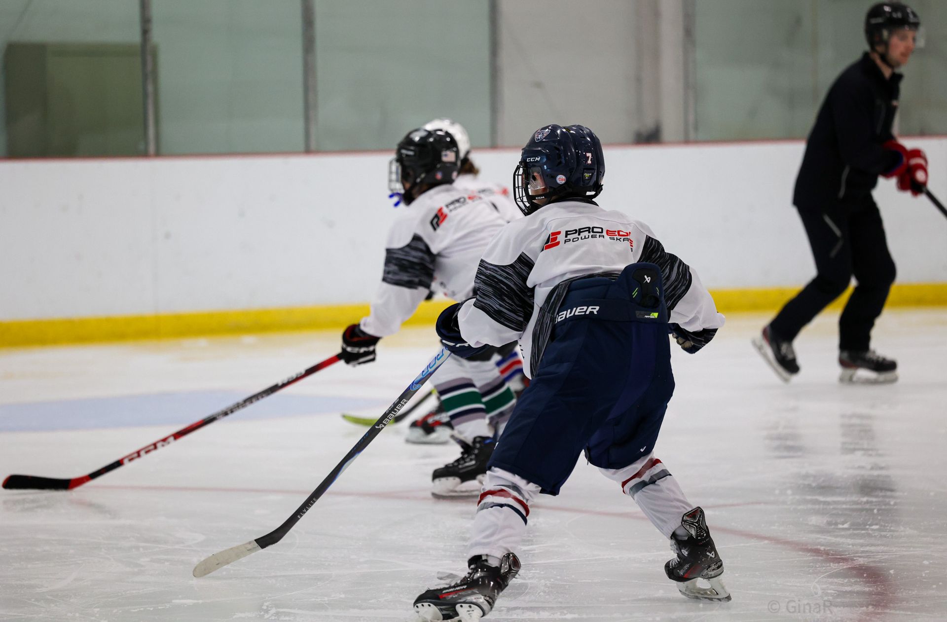 Youth hockey players skating after the puck on an indoor ice rink with a coach in the background