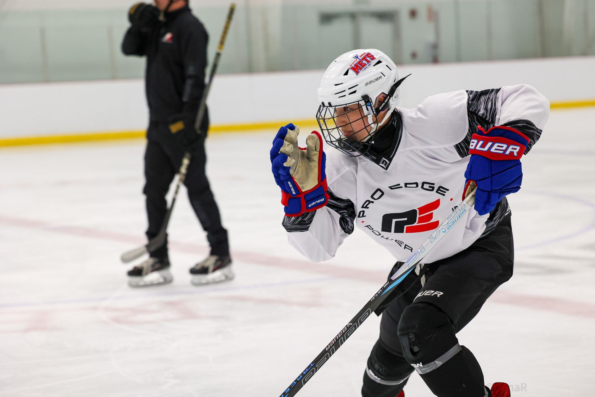 Hockey player in white jersey skating on ice, gripping stick, with a coach blurred in the background