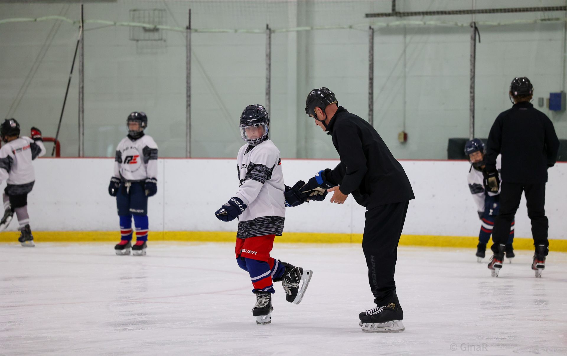 Youth ice skater practicing with coach on an indoor rink, both wearing helmets.