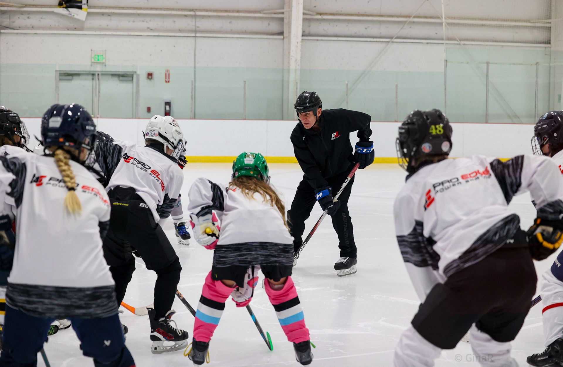 Youth hockey players in white jerseys face a coach on an indoor ice rink.
