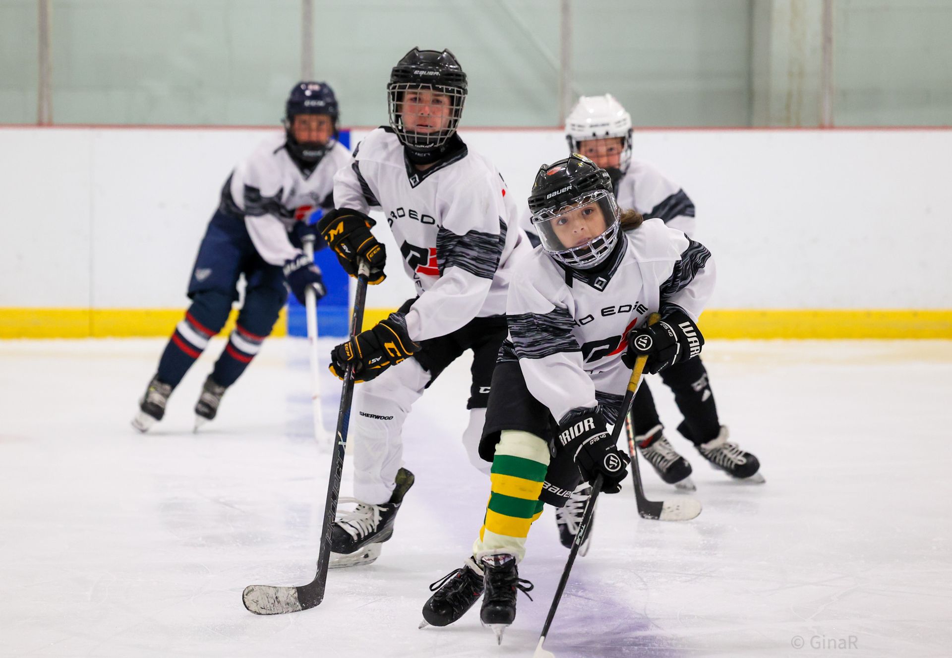 Youth ice hockey players racing on the rink, one in white and green leading with the puck.