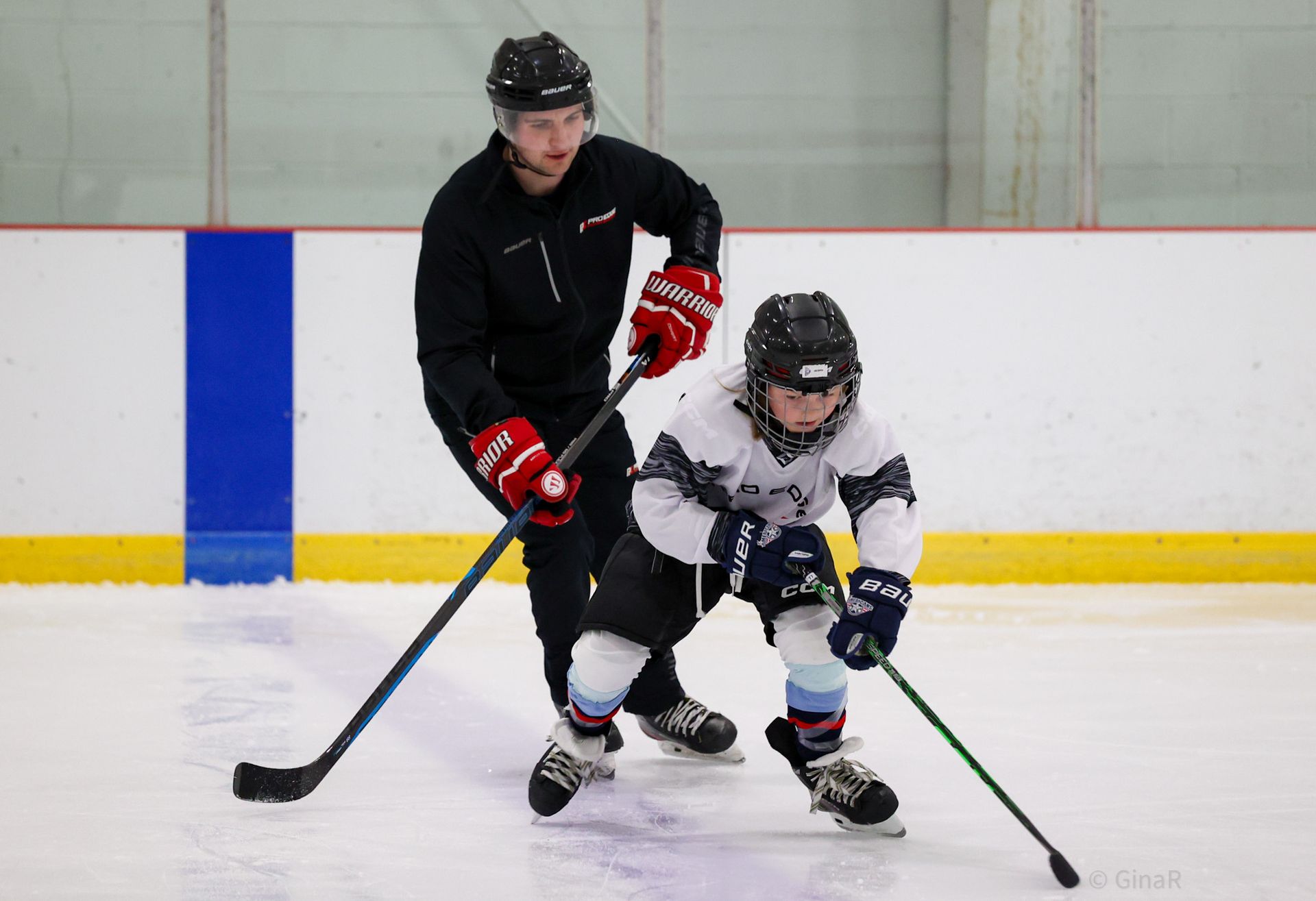 Two hockey players in black and white gear skating on an indoor ice rink with sticks and helmets