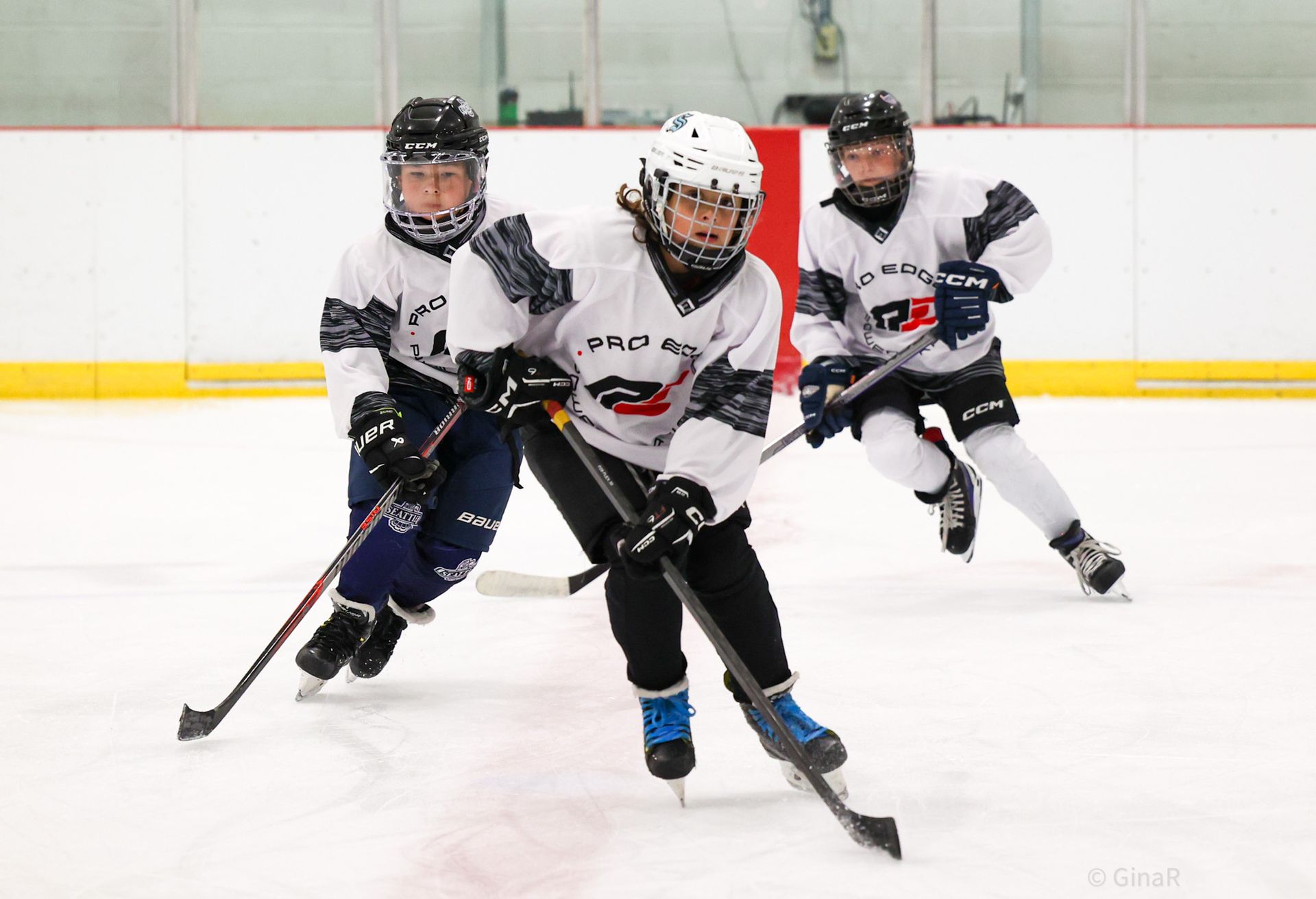 Three youth hockey players in white uniforms skating fast on an ice rink