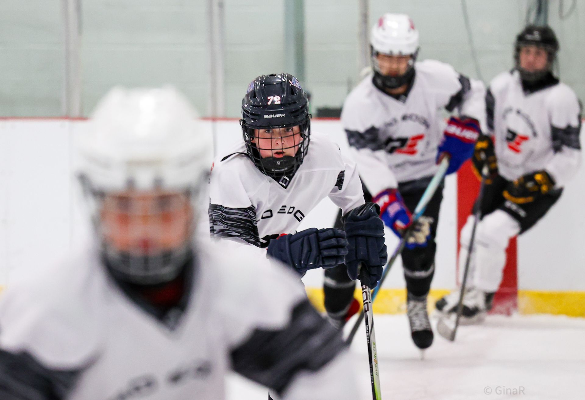 Youth hockey players in white jerseys skating on an indoor rink, one centered and focused in the foreground