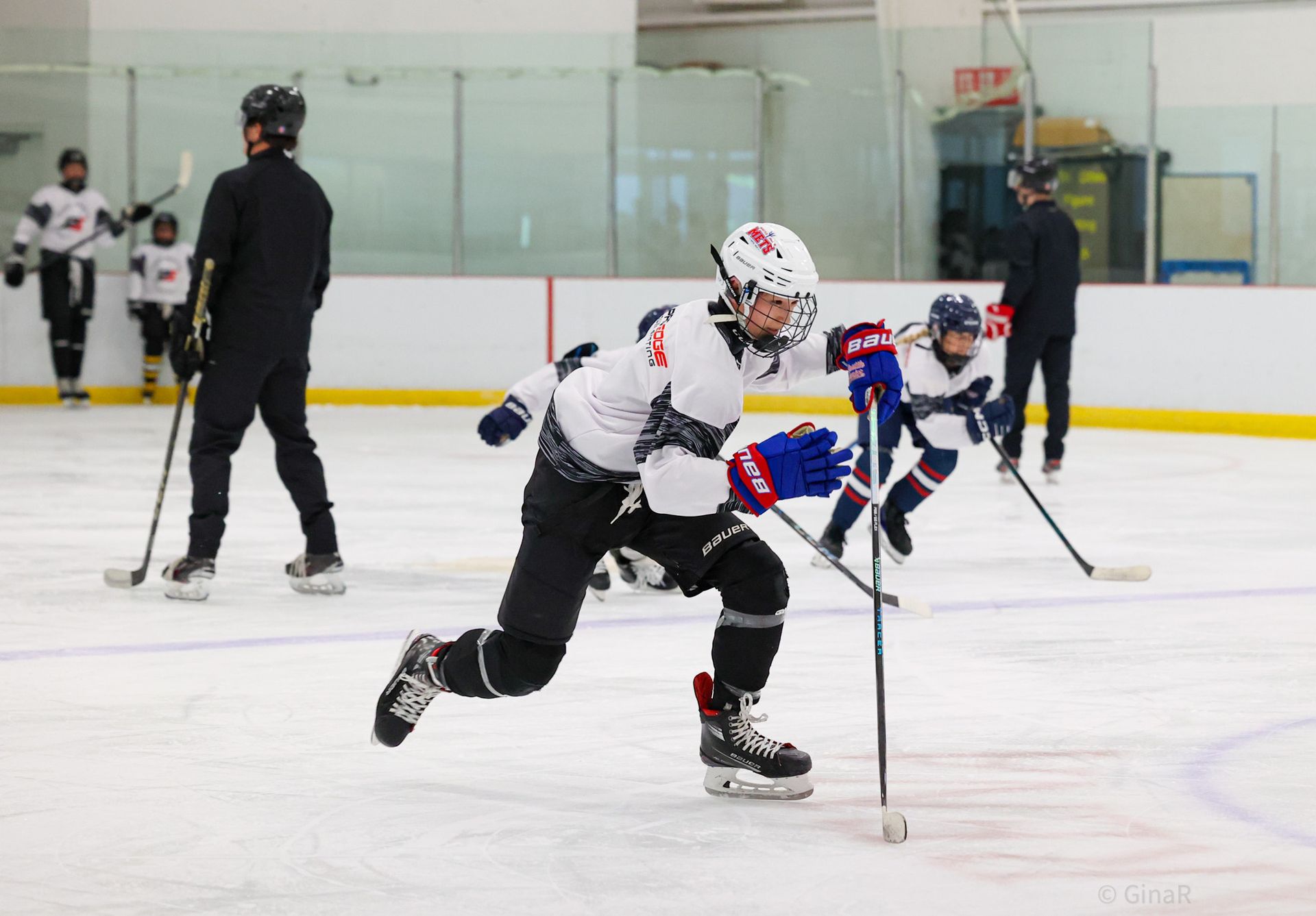 Two hockey players sprint on an indoor ice rink, one in white and red leading with the puck.