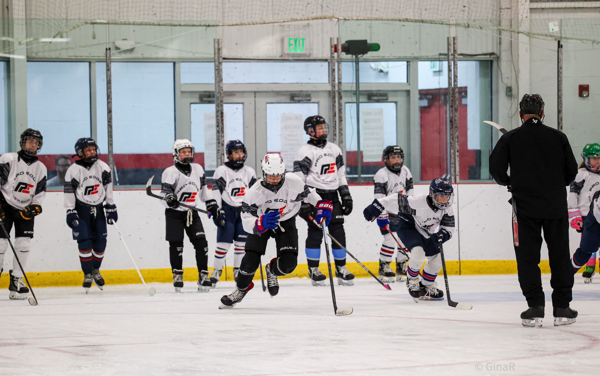 Youth hockey players in white jerseys line up on an indoor ice rink with a coach nearby.