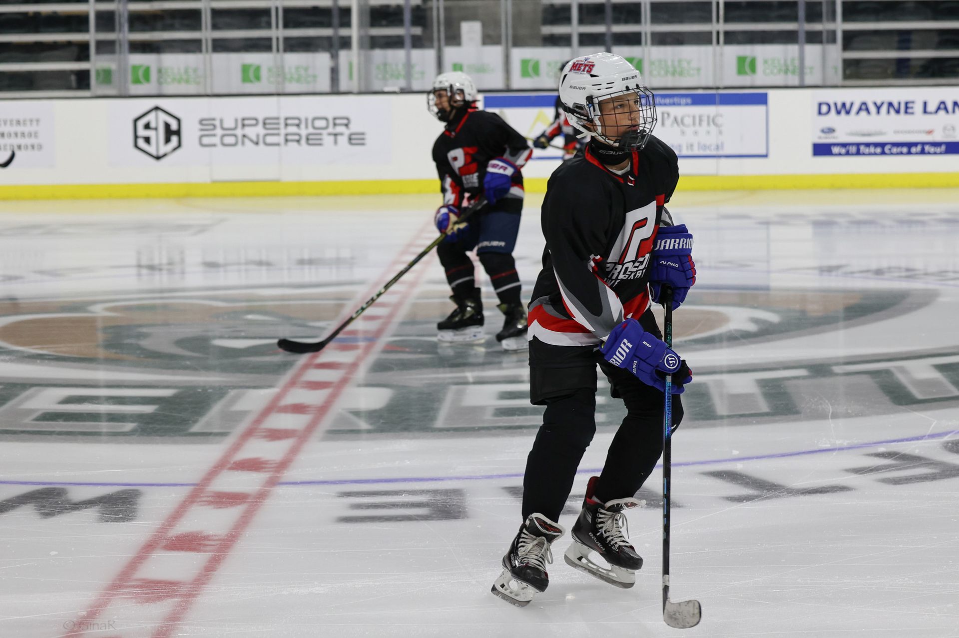 Two hockey players in black jerseys skate on an indoor ice rink, holding sticks near center ice.