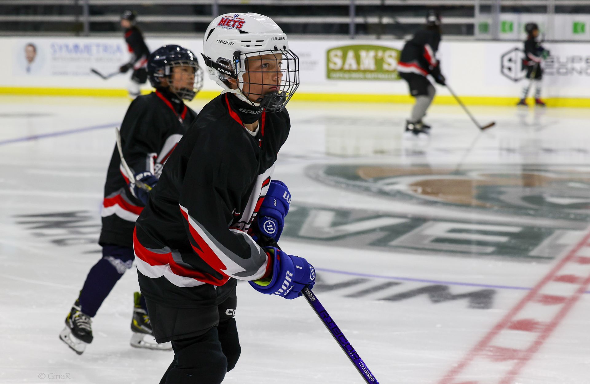 Youth hockey players skating on an ice rink, with one in black and red in the foreground holding a blue stick.