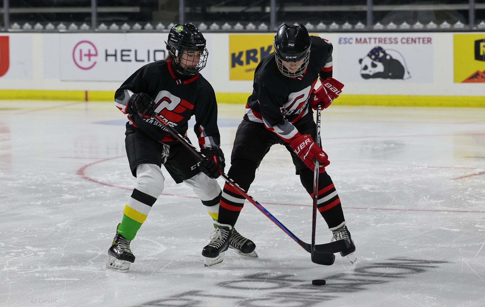 Two hockey players in black and red uniforms compete for the puck on an ice rink.