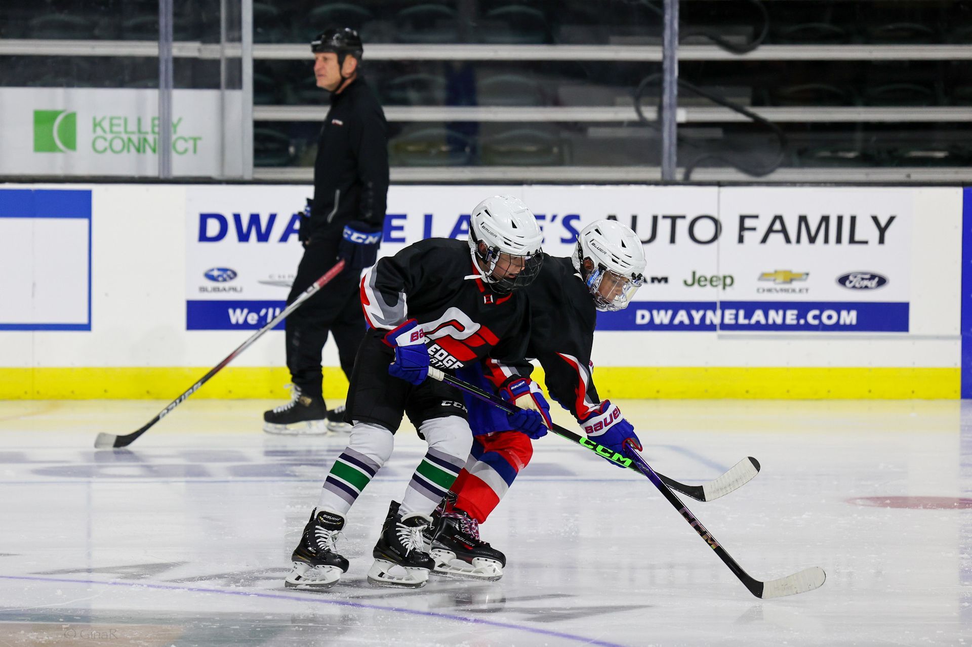 Two hockey players in red and black battle for the puck on an ice rink, with a referee nearby.