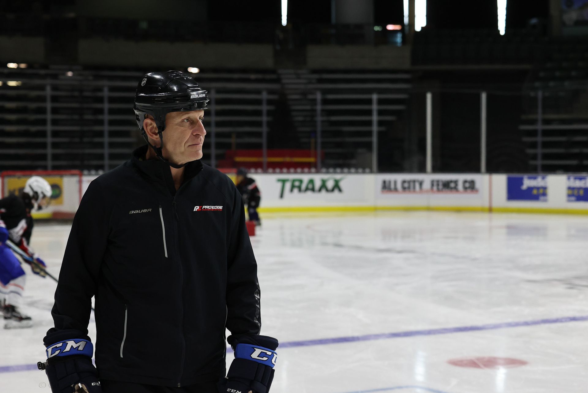 Hockey coach in black gear stands on the ice rink with players practicing in the background