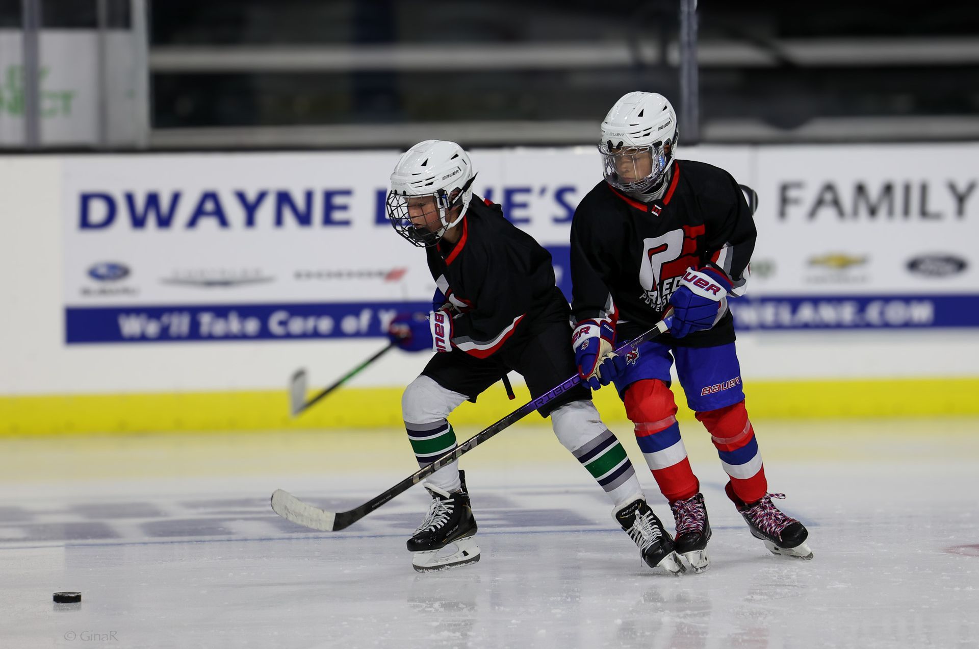Two hockey players in black and blue uniforms chase the puck on an ice rink.