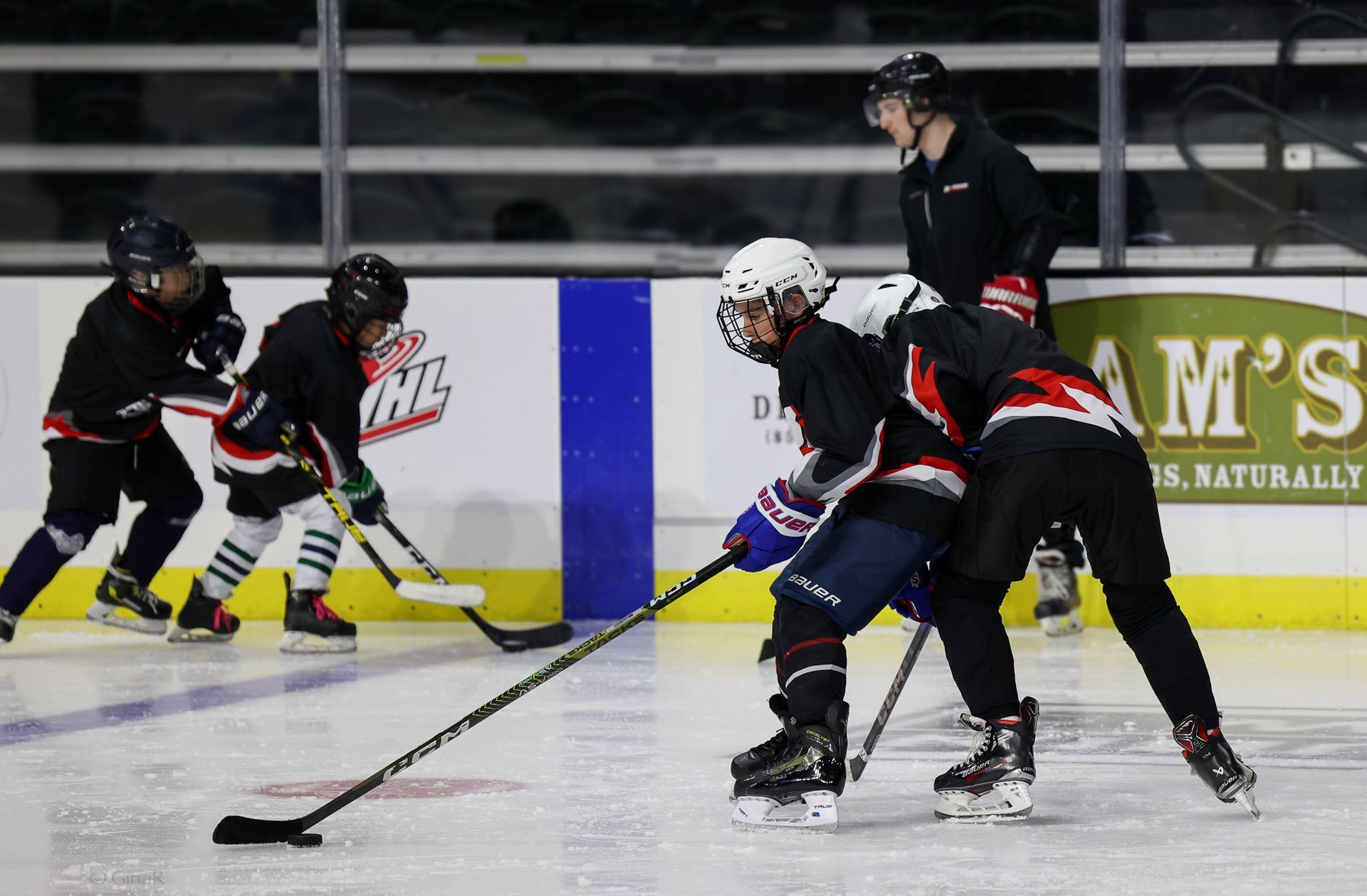 Ice hockey players in black uniforms chase the puck on the rink near the boards.