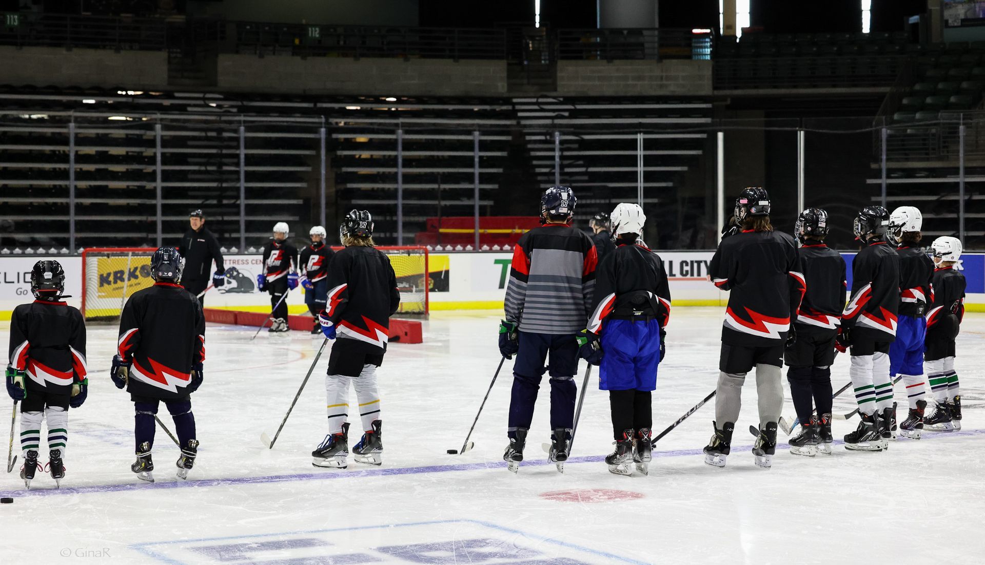 Youth hockey players in black and blue jerseys gathered on the ice during practice at an indoor rink