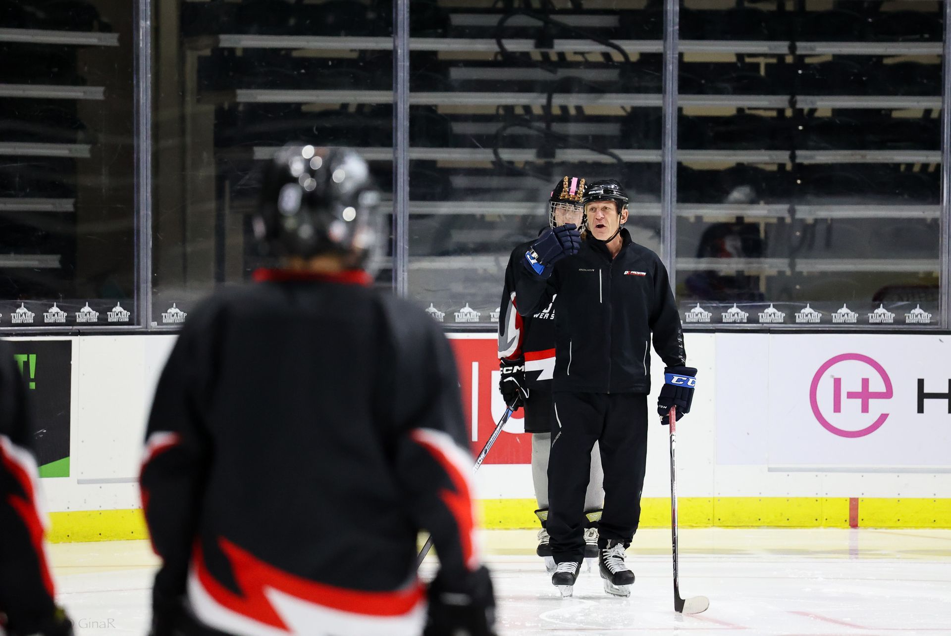 Ice hockey coach in black gear on rink, speaking to a player during practice.