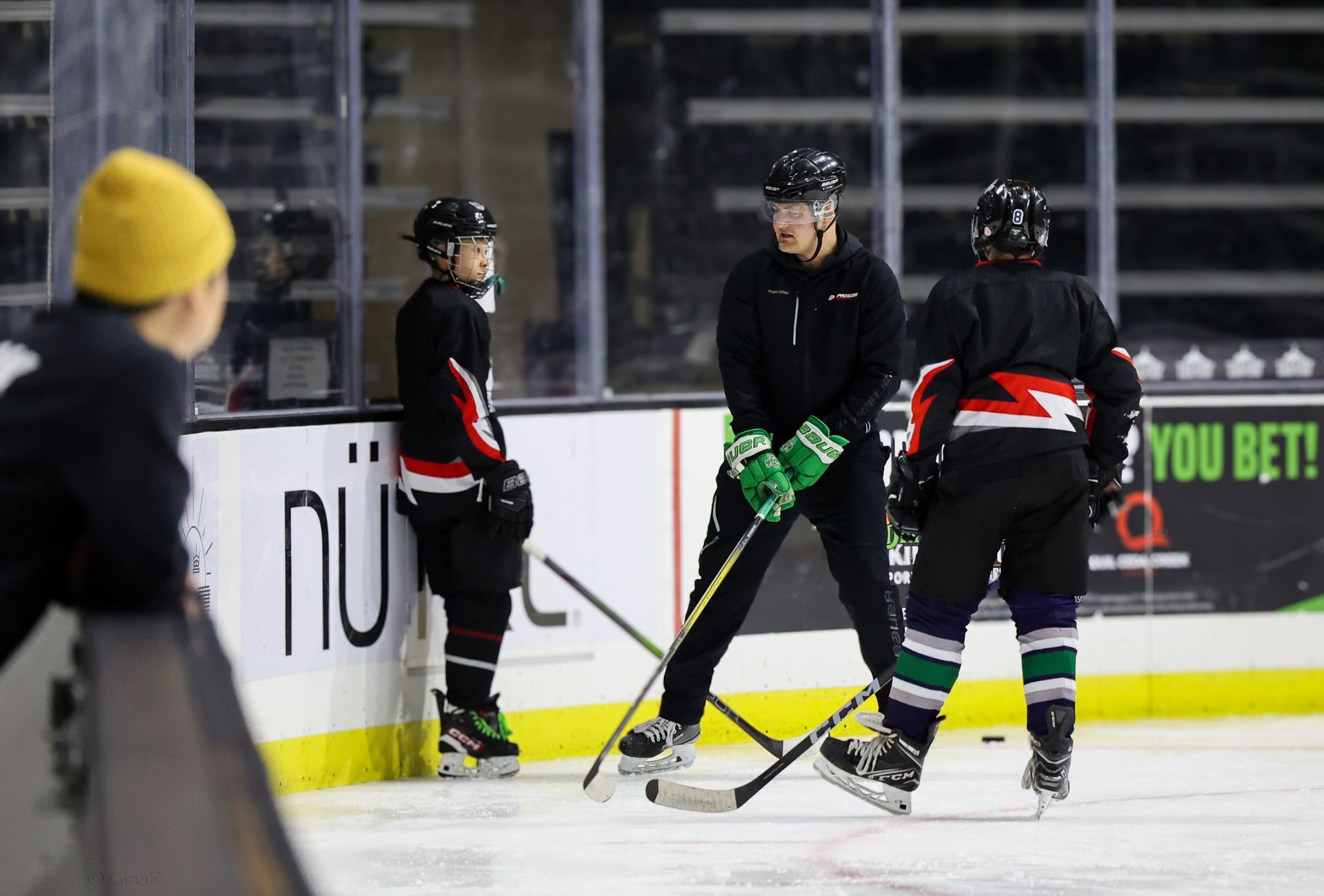 Hockey players in black uniforms gather on the ice during practice near the boards.