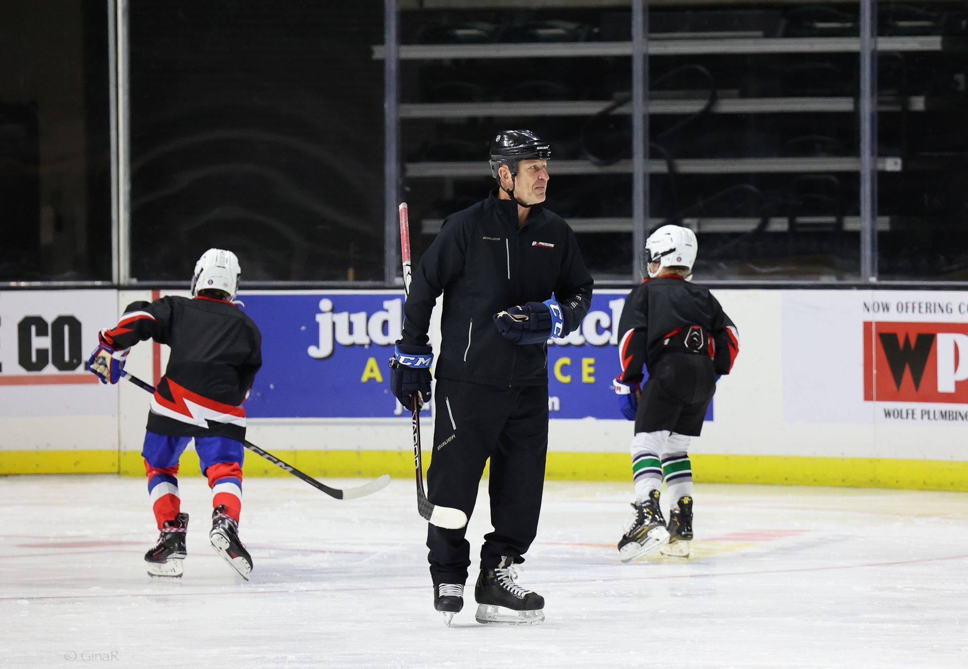 Ice hockey coach in black stands on the rink while two young skaters in helmets glide nearby.