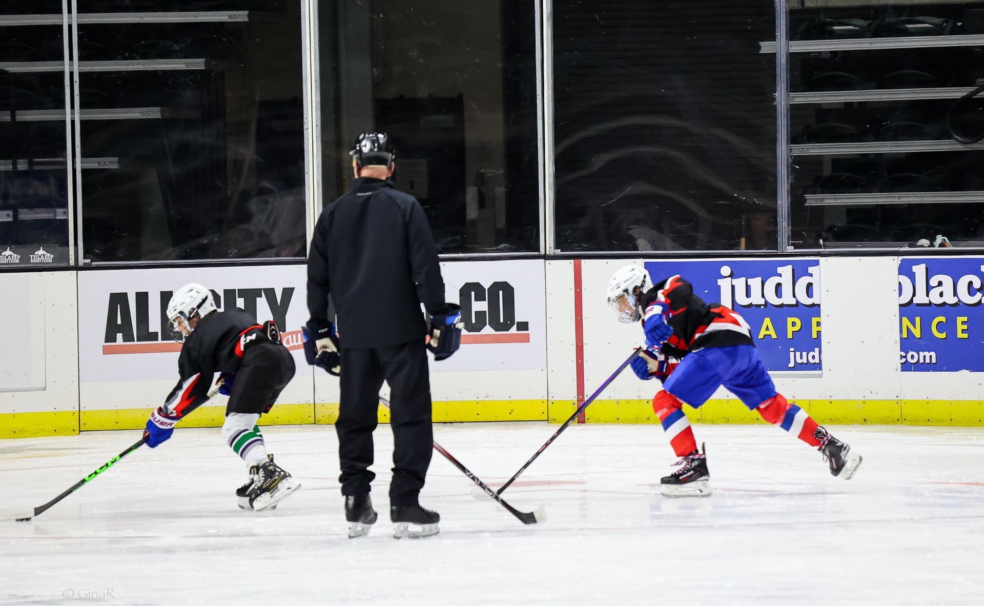 Ice hockey players in blue and red crouch for a faceoff near the boards, with a referee between them.