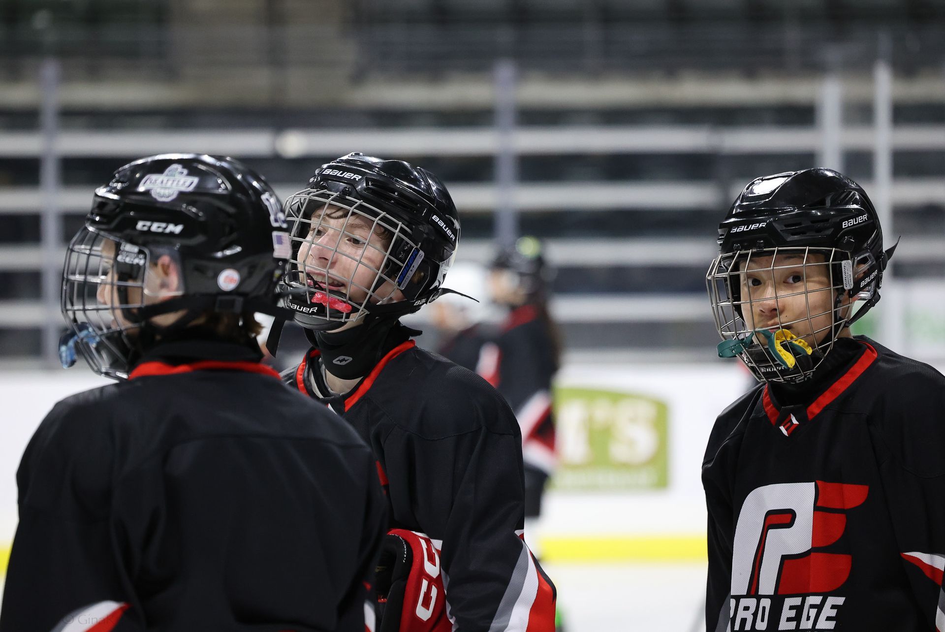 Three hockey players in black jerseys and helmets stand on the ice, one facing camera with a chin guard.