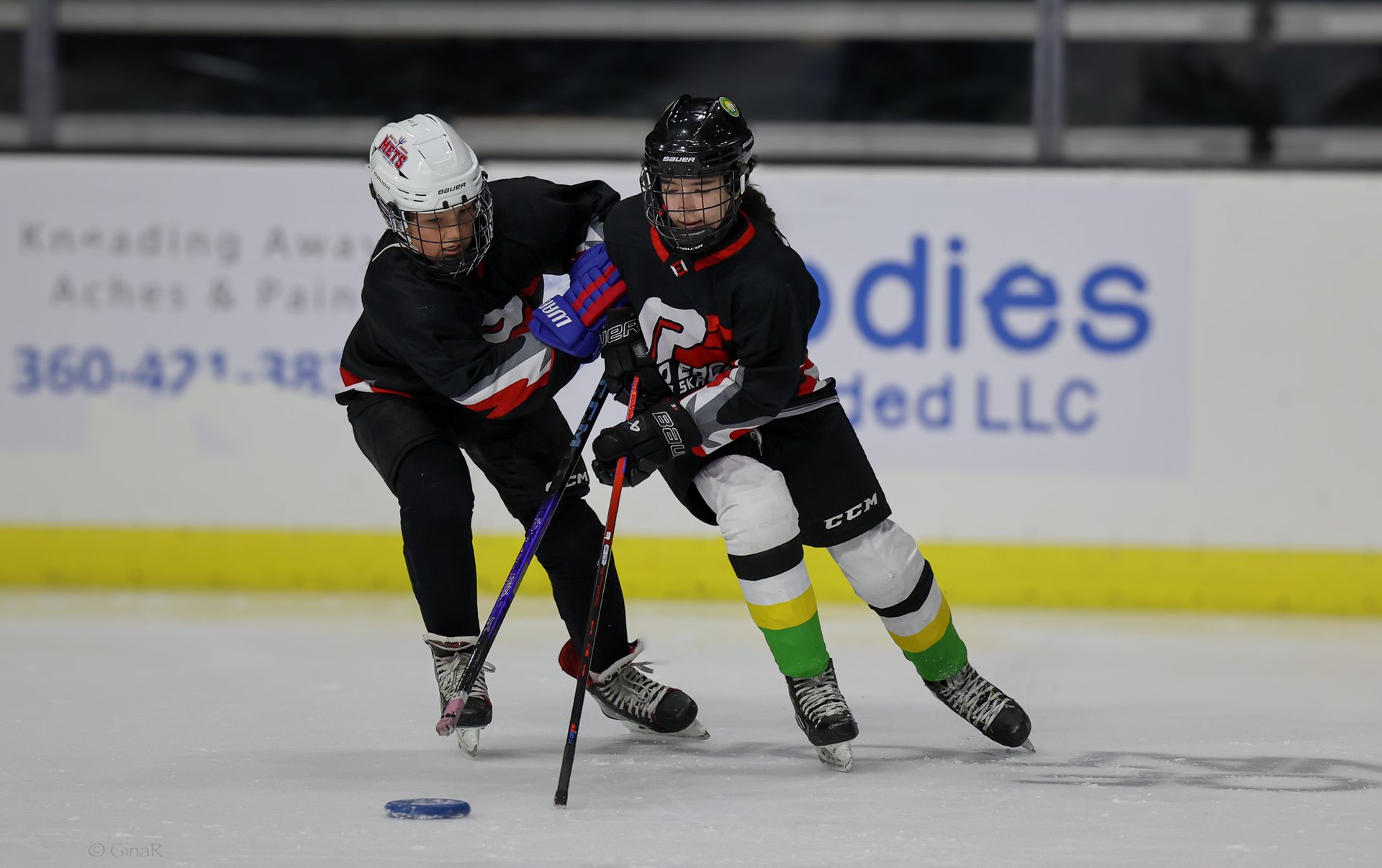 Two hockey players in black uniforms race for the puck on the ice near the boards.