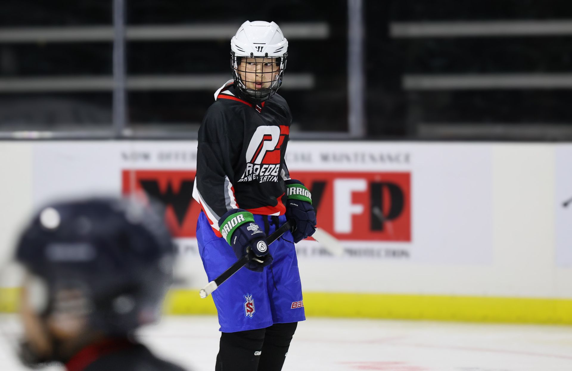 Hockey player in black jersey and blue pants stands on the ice, holding a stick and looking ahead.