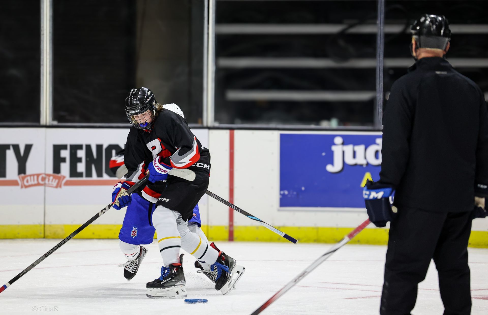 Two hockey players battling for the puck on an indoor ice rink, with a referee nearby.