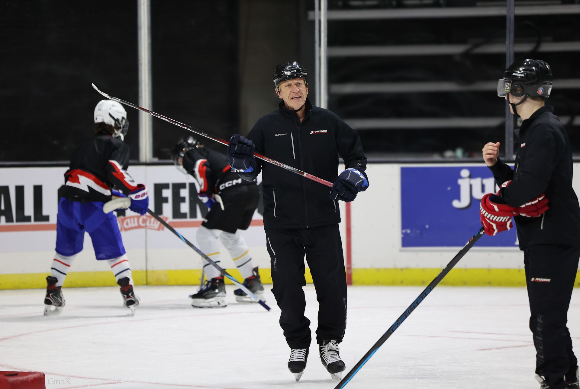 Hockey coach on the ice instructing two young players during practice, with hockey sticks and rink boards visible