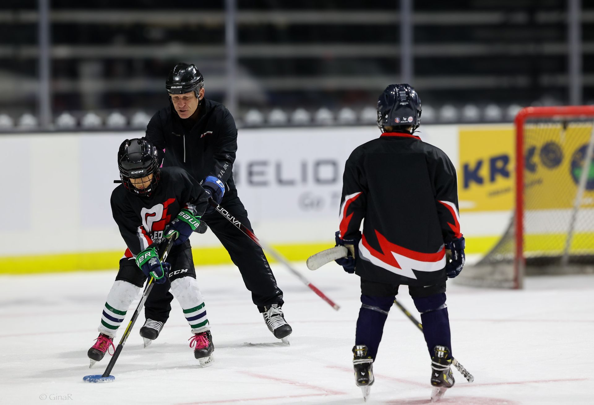 Youth hockey players skating on an indoor rink, with a coach behind them and the goal visible.
