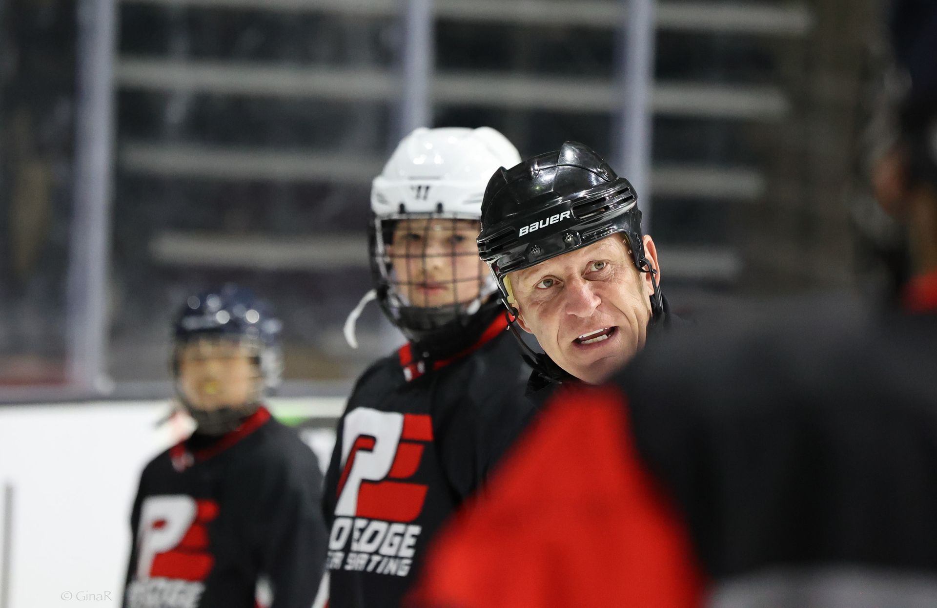 Hockey coach in black helmet and red-and-black jersey watching play on the ice