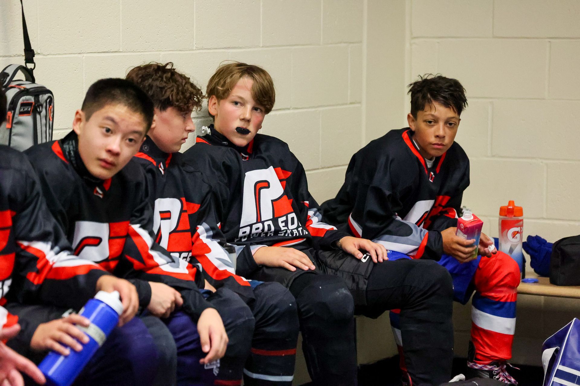 Young hockey players in black jerseys sit on a bench, holding water bottles and resting during a game.