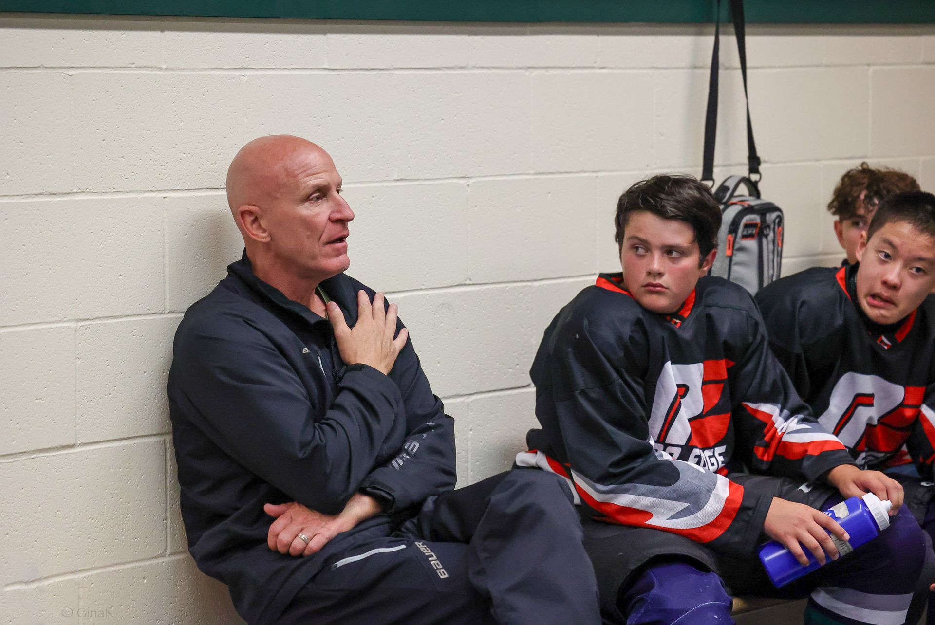 Coach talking with two hockey players in a locker room, one hand on his chest, players seated on bench
