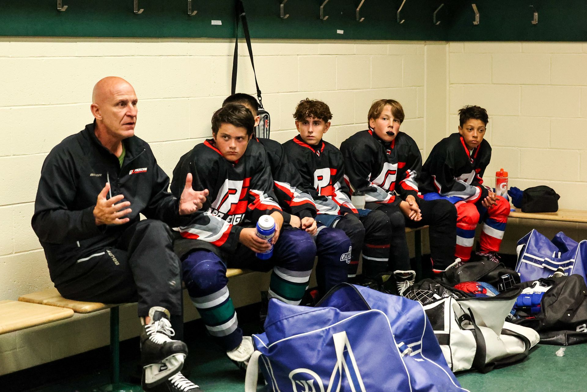 Youth hockey players and coach sitting on a bench in the locker room, wearing black, red, and blue uniforms.