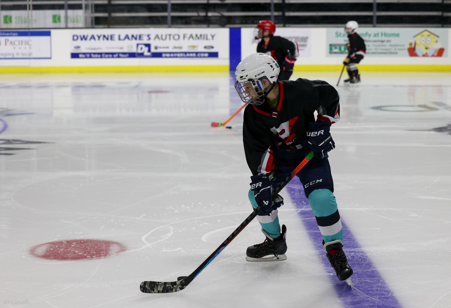Youth ice hockey player in black jersey handling the puck on an indoor rink, teammate and goalie in background