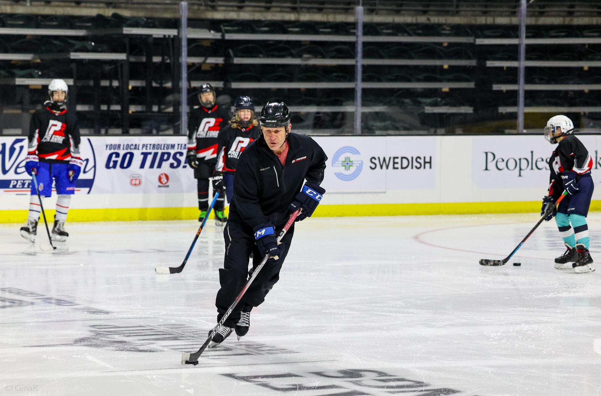 Ice hockey players skating during practice on an indoor rink, with a coach in black in the foreground