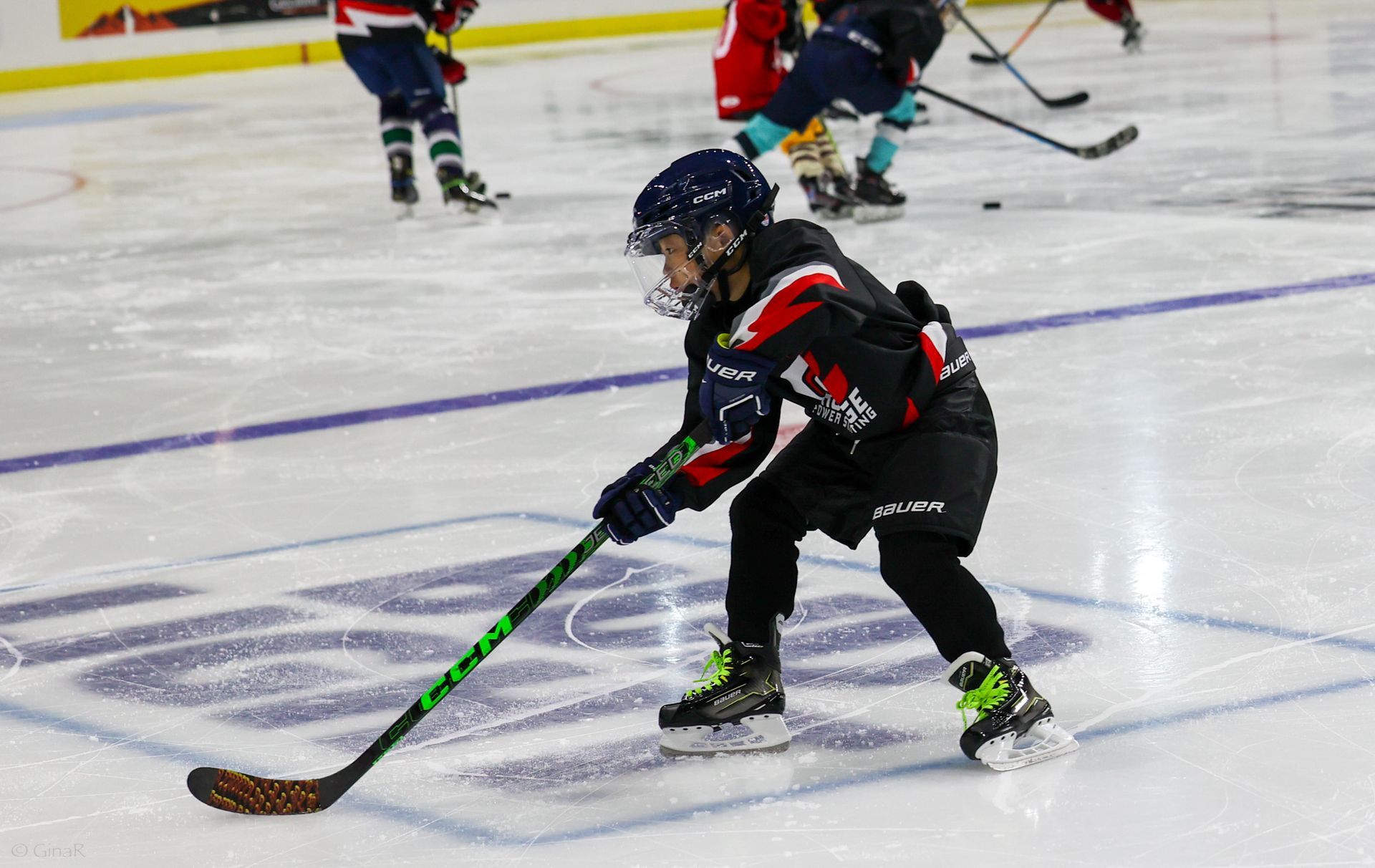 Youth hockey player in black and red skating on ice, stick extended toward the puck.
