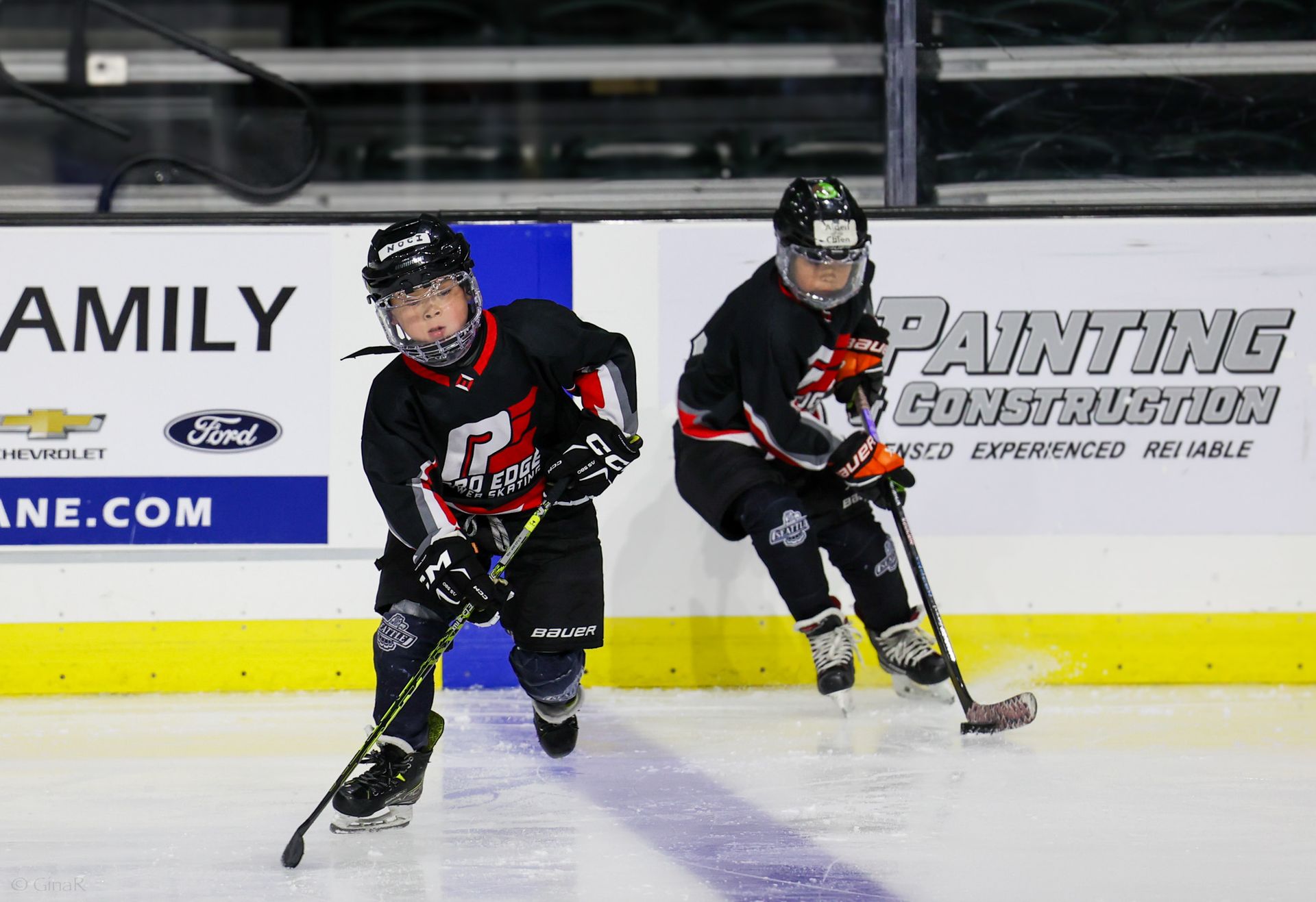 Two hockey players in black uniforms skating on ice during a game, one leading the puck