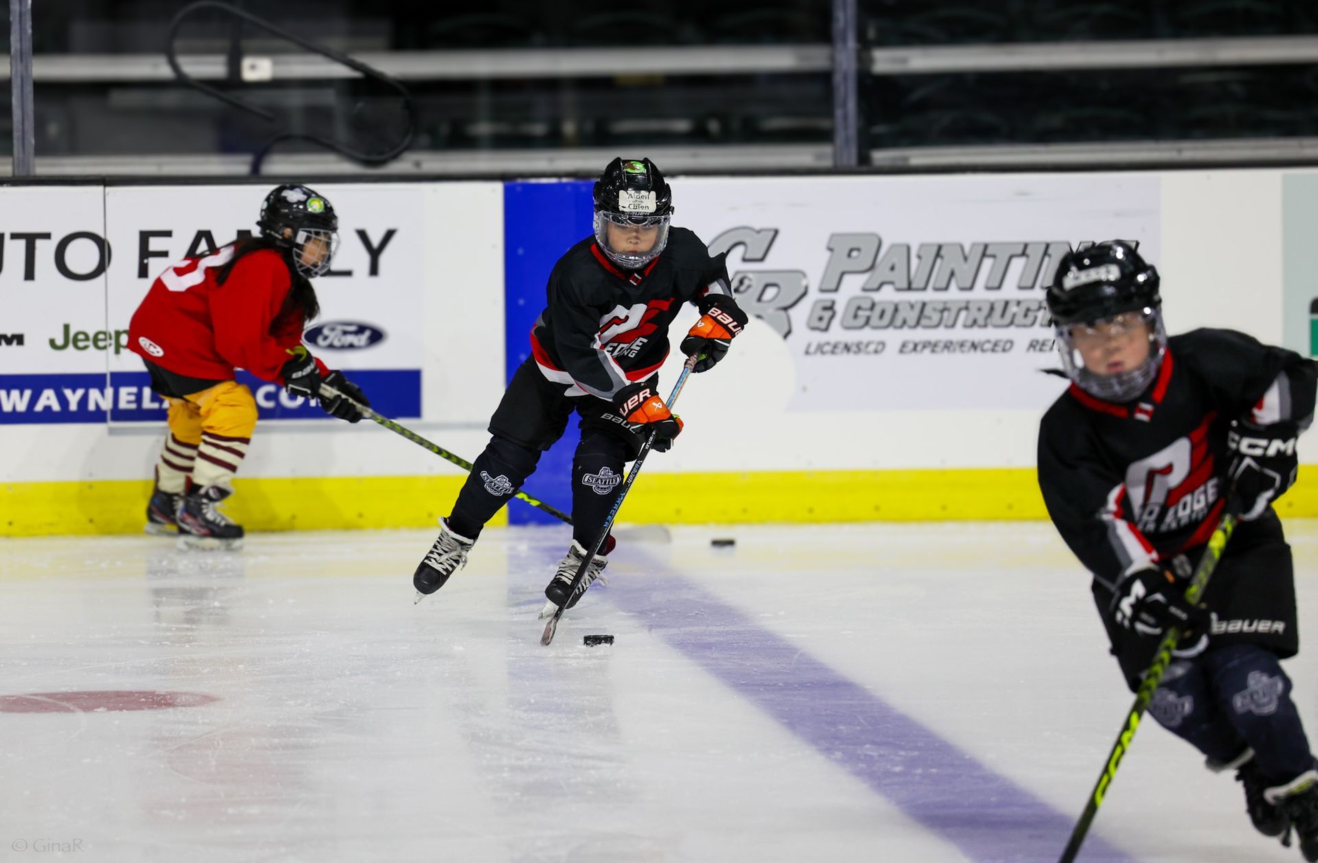 Youth hockey players in black and red skate on an indoor rink during a game.