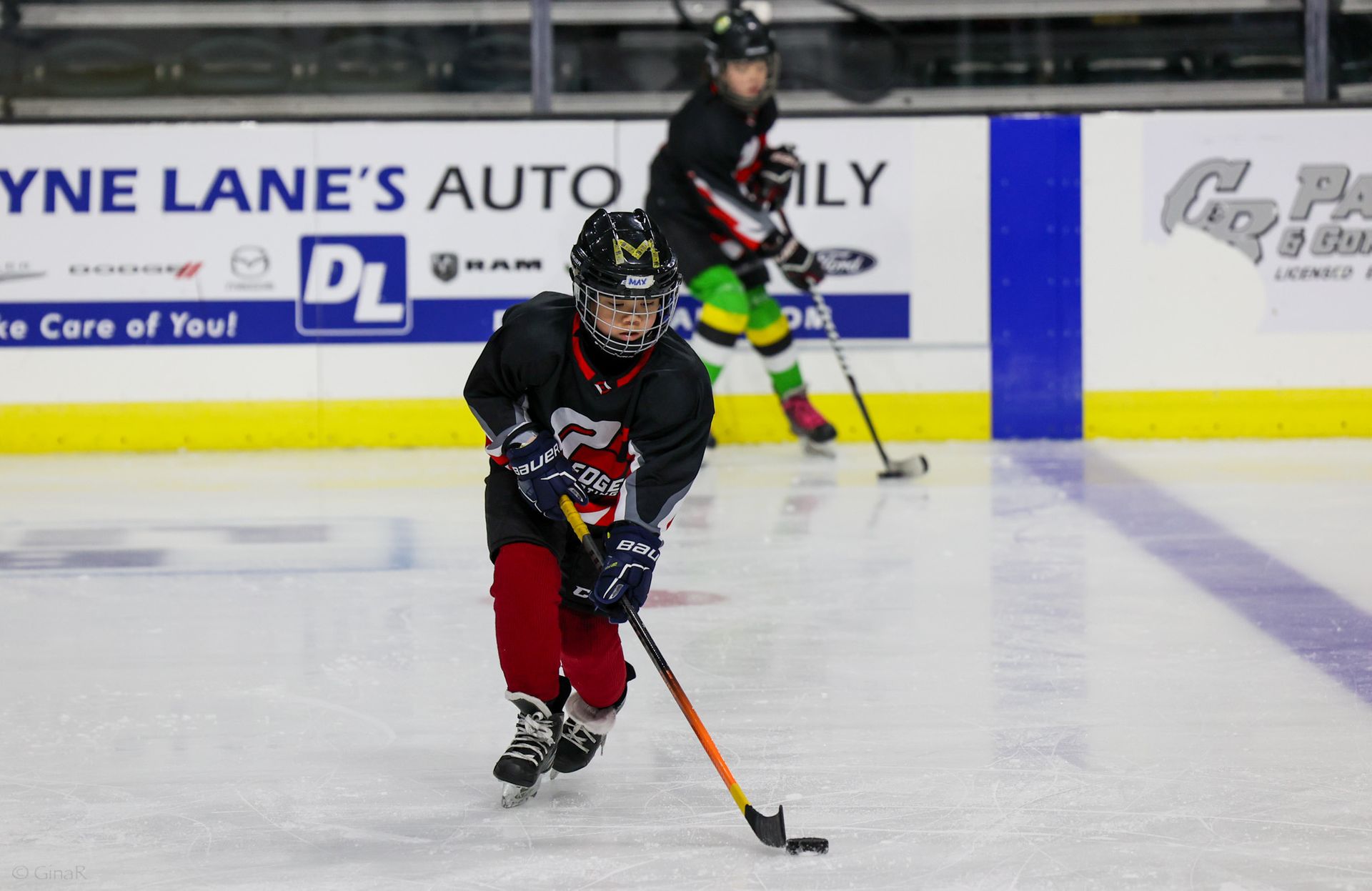 Youth hockey player in black and red skates with puck on ice, another player behind, rink boards in background