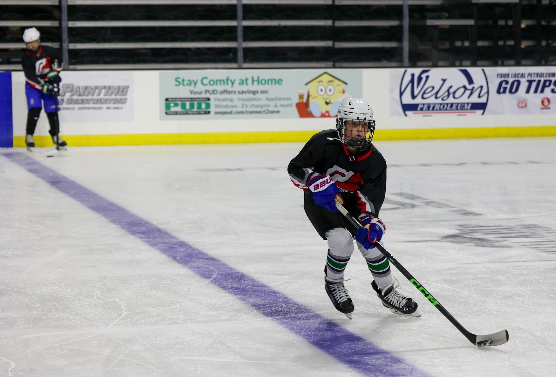 Hockey player in black skates across the ice, stick on the puck, with another player in the background.