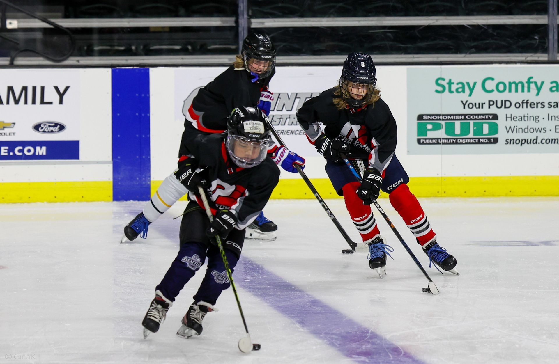 Youth hockey players in black jerseys race for the puck on an indoor ice rink.