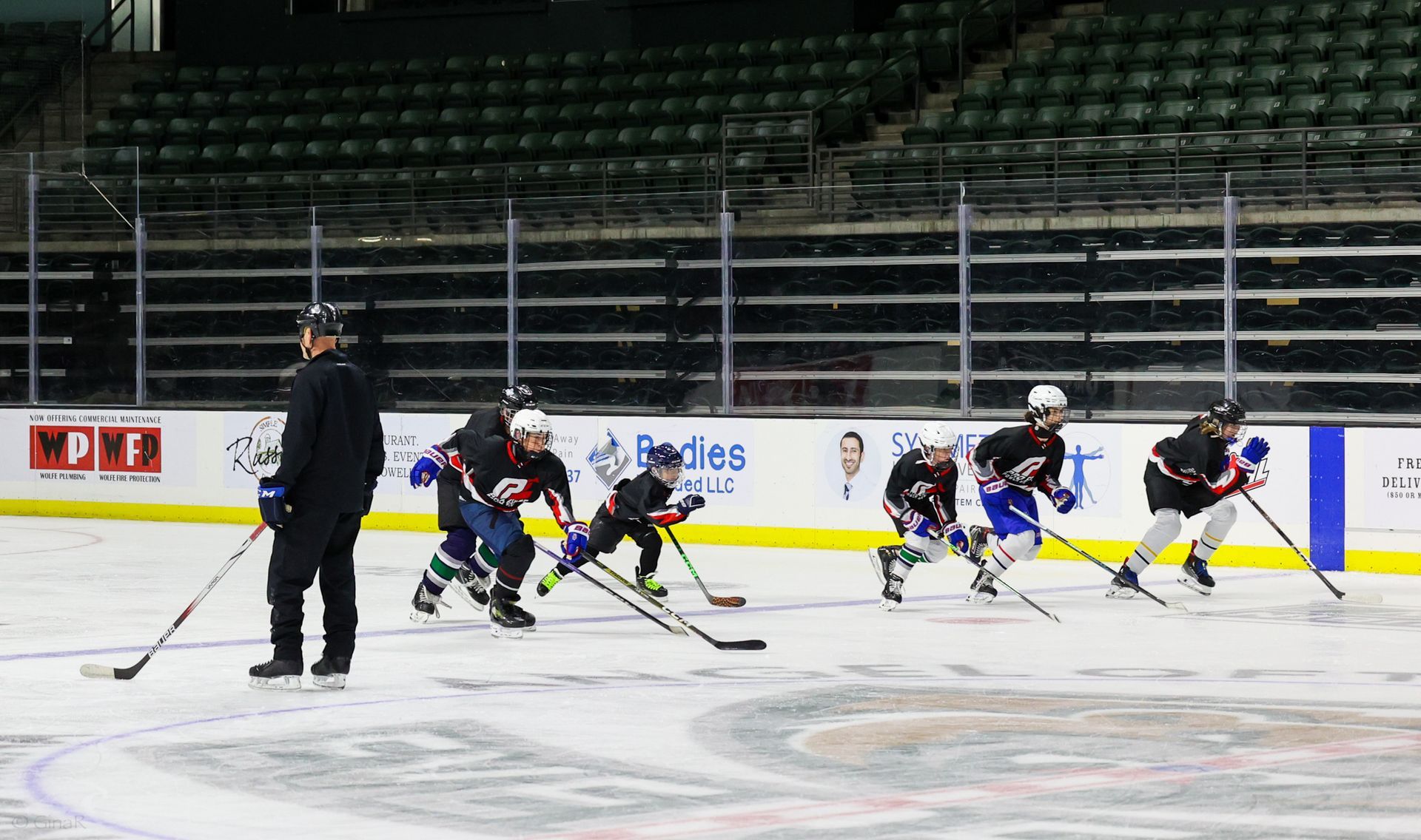 Ice hockey practice on an indoor rink, with players skating and a coach watching from the left.