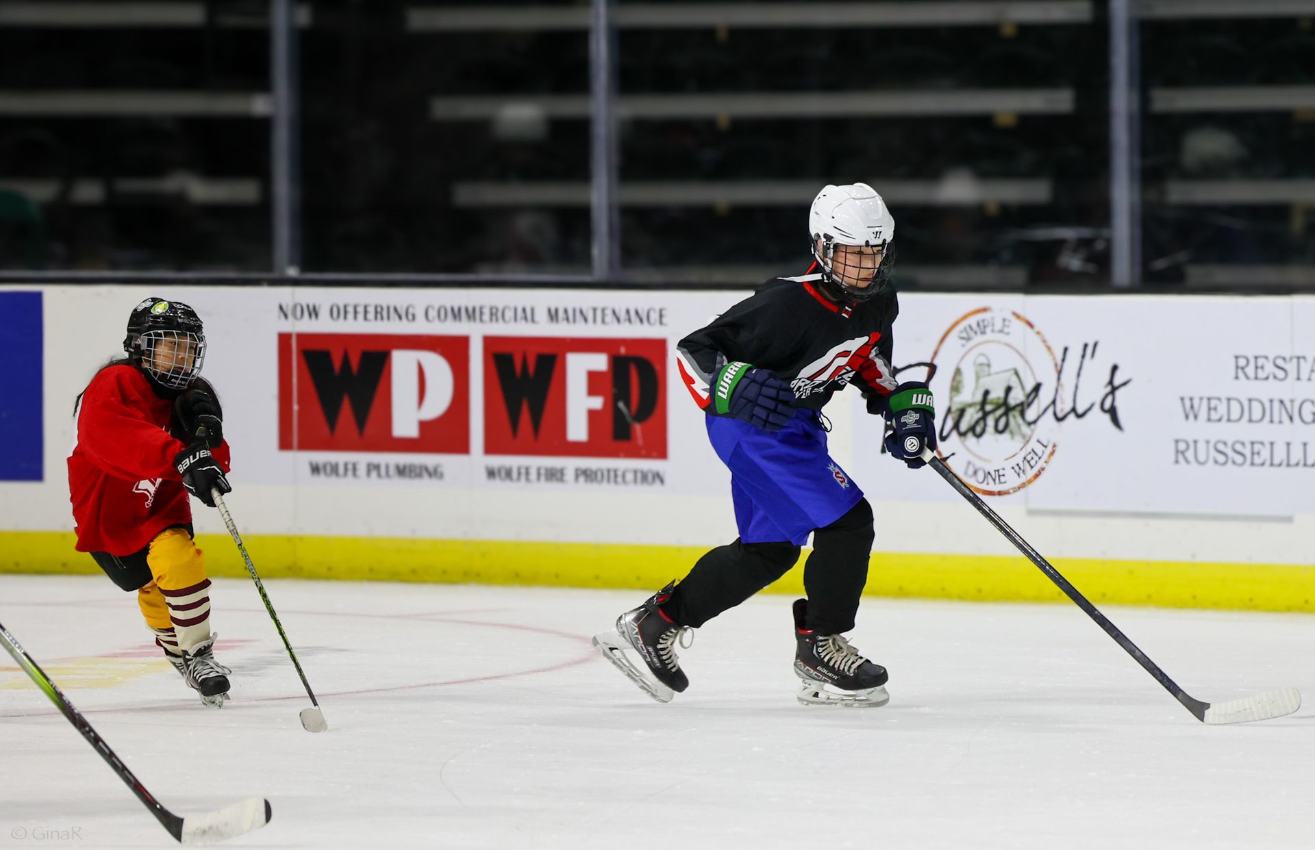 Two hockey players race on an indoor rink, one in red chasing a player in blue and white.