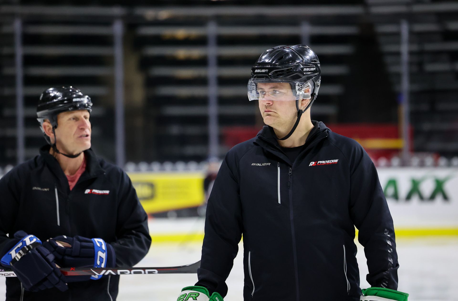 Two hockey players in black jackets and helmets stand on an ice rink during practice.